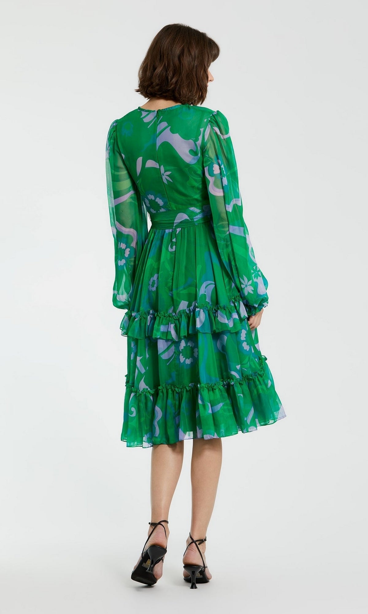 A woman faces away, wearing the Short Wedding Guest Dress 68760 by Mac Duggal—a green floral knee-length a-line dress with ruffles, long sleeves, delicate tiers—and black strappy heels, set against a plain background.
